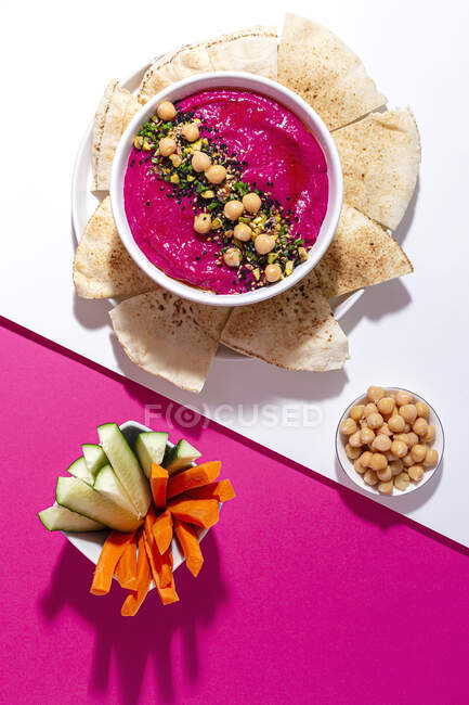 Top view of appetizing beetroot hummus garnished with chickpea served on two colored background with bread and fresh carrot and cucumber sticks — Stock Photo