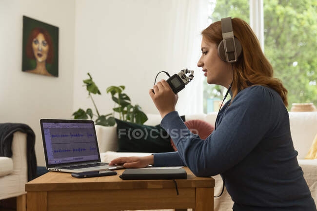 Side view of female in headphones recording podcast while speaking in microphone and using laptop at home — Stock Photo