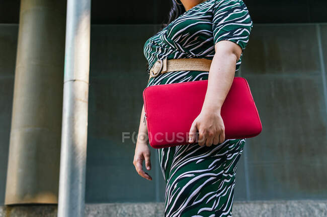 Side view of anonymous cheerful young female in stylish striped outfit with red laptop case in hands smiling while standing against contemporary city building — Stock Photo