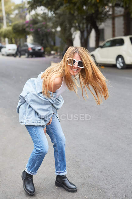 Jeune femme élégante en lunettes de soleil et vêtements en denim riant dans la rue tout en profitant week-end et en regardant la caméra — Photo de stock