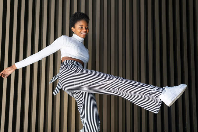 Baixo ângulo de otimista preto fêmea em roupas elegantes sorrindo e balançando a perna enquanto dança contra a parede listrada na rua da cidade — Fotografia de Stock