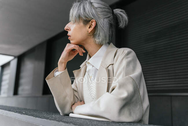 Serious transgender female in trendy coat leaning on gray fence while standing in street looking away — Stock Photo
