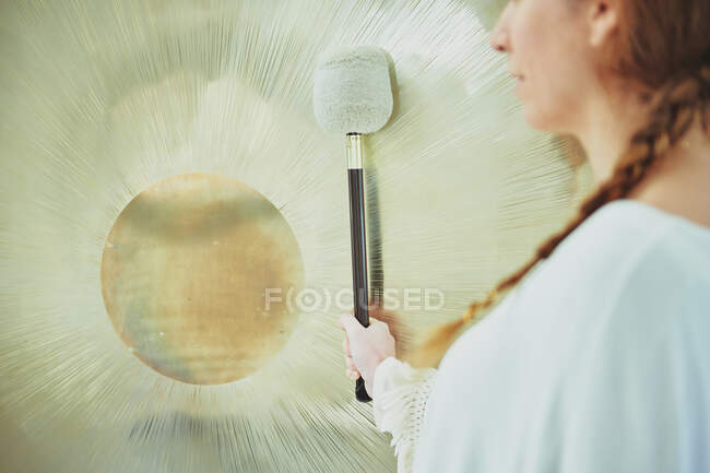 Side view of crop adult female in white clothes with mallet playing suspended gong during spiritual practice — Stock Photo