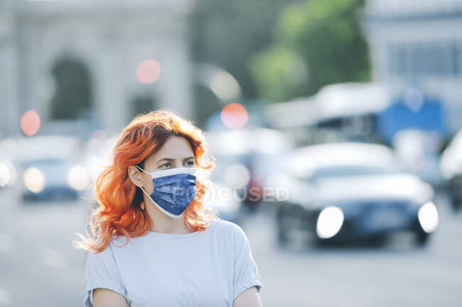 Mujer con cabello rojo con máscara médica protectora en la calle de la ciudad durante la epidemia de coronavirus - foto de stock