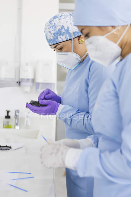 Vista lateral de las mujeres de los cultivos médicos en uniformes y máscaras respiratorias en la mesa con herramientas profesionales en la clínica - foto de stock