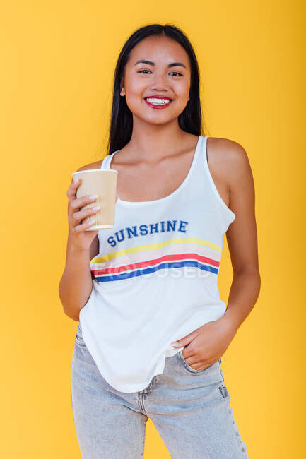 Encantada Asiática fêmea em roupa de verão em pé com tafetá xícara de café no fundo amarelo e olhando para a câmera — Fotografia de Stock