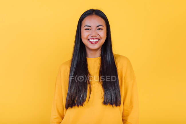 Encantada Asiática fêmea em pé no fundo amarelo no estúdio enquanto olha para a câmera — Fotografia de Stock
