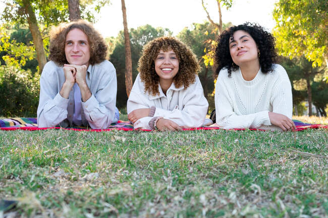Diverse male and female friends with curly hair lying on colorful blanket on meadow and talking in summer — Stock Photo