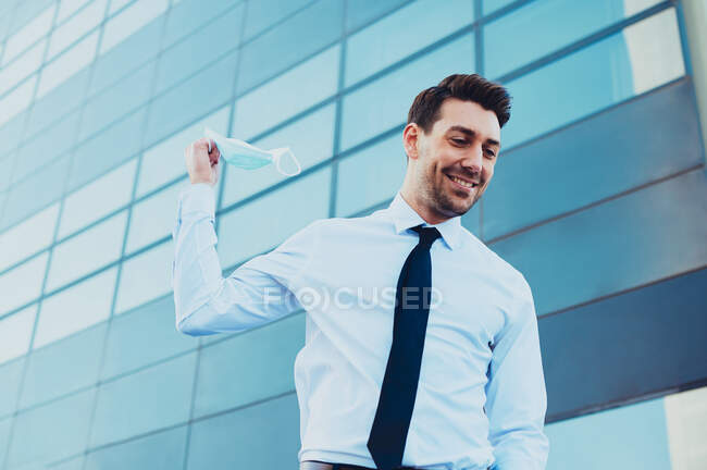From below of content male entrepreneur in formal wear holding medical mask looking away in town during coronavirus pandemic — Stock Photo