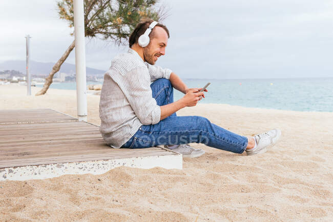 Вид сбоку веселого молодого бородатого мужчины в беспроводных наушниках, слушающего музыку и просматривающего мобильный телефон, отдыхающего в одиночестве на песчаном пляже в летний день — стоковое фото
