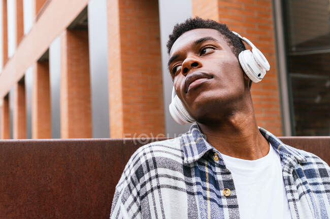 Unbeweglicher Afroamerikaner, der mit kabellosen Kopfhörern in der Stadt steht und wegschaut — Stockfoto