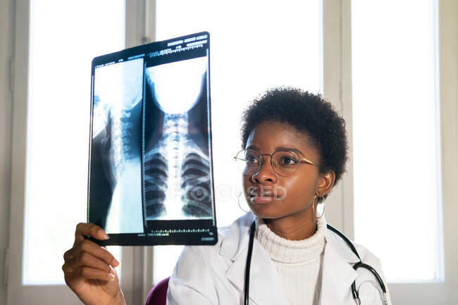 Giovane medico donna nera in piedi vicino alla finestra ed esaminando la scansione a raggi X mentre si lavora in clinica — Foto stock