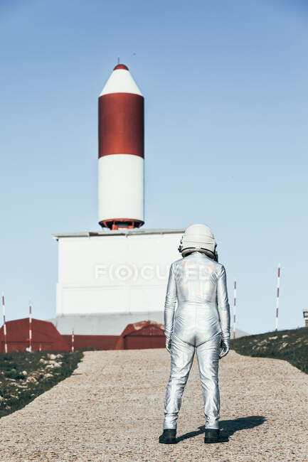 Voltar ver o homem no traje espacial em pé no chão rochoso contra as antenas em forma de foguete listrado no dia ensolarado — Fotografia de Stock