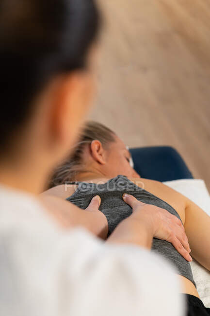 Сверху обрезается неузнаваемая массажистка, улыбающаяся и массирующая спину женщине во время работы в физиотерапевтической клинике — стоковое фото
