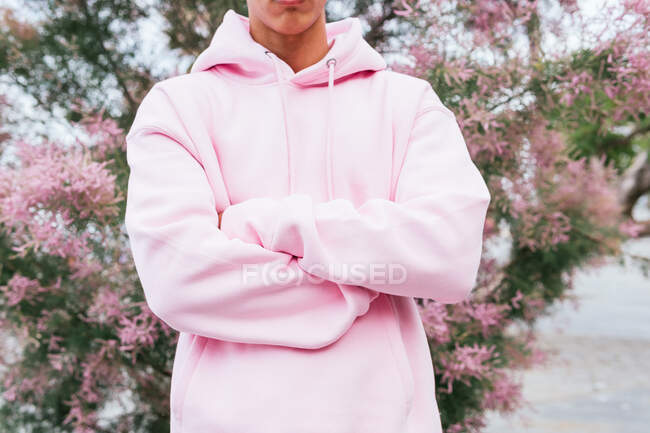 Cortado irreconhecível homem étnico com penteado afro tingido vestido com capuz rosa na moda de pé com os braços cruzados contra a árvore florescente e olhando para a câmera — Fotografia de Stock