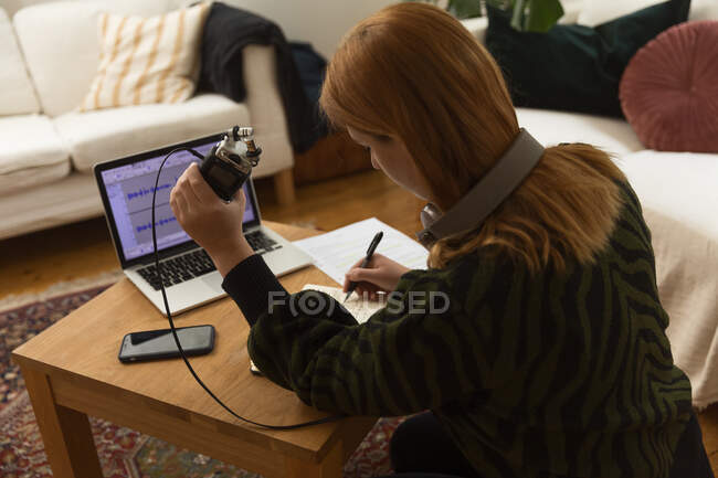 Vista lateral del anfitrión de radio femenino enfocado con micrófono y auriculares escribiendo en el bloc de notas mientras se prepara para grabar podcast en casa - foto de stock