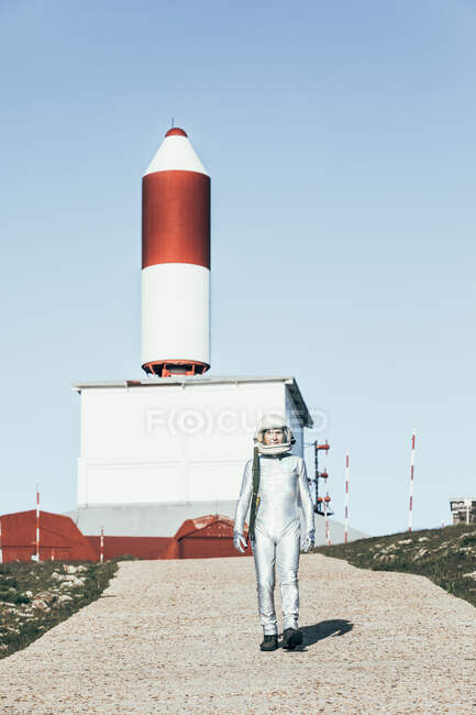 Человек в скафандре, стоящий на скалистой земле против полосатых антенн в форме ракеты в солнечный день — стоковое фото