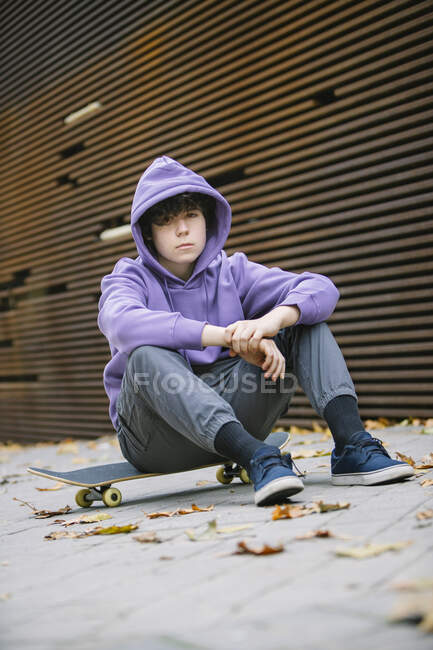 Full body of teenage boy in casual clothes with hoodie looking at camera while sitting on skateboard at wall on street with fallen leaves — Stock Photo