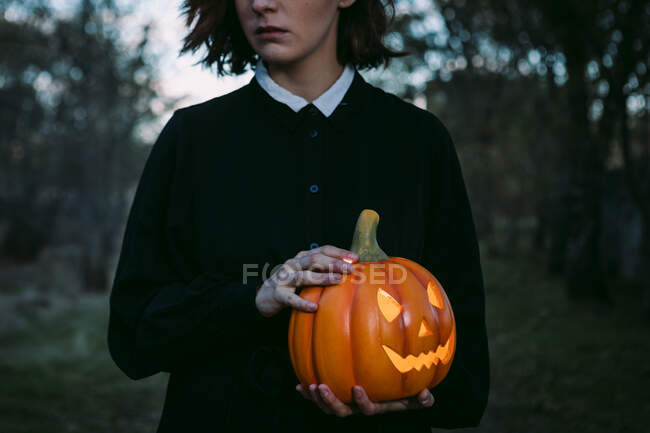 Cropped unrecognizable female in black dress standing with glowing pumpkin lantern in dark woods on Halloween — Stock Photo