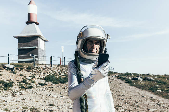 Hombre de edad en traje espacial y datos de navegación casco en el teléfono inteligente, mientras que de pie cerca de edificios industriales con antenas en forma de cohete - foto de stock