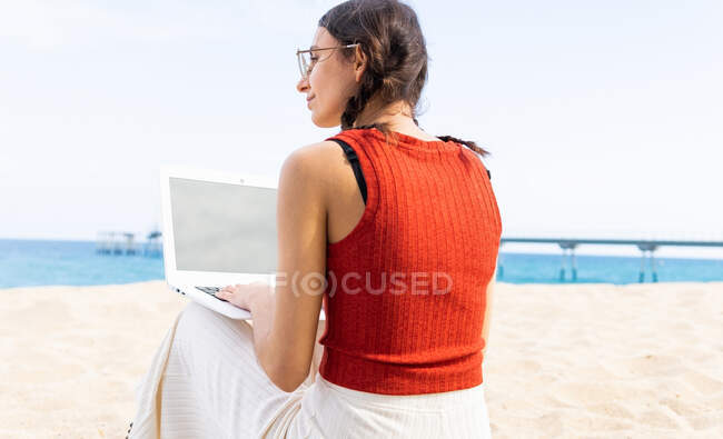 Vista posterior del contenido freelancer femenina sentada en la orilla del mar y escribiendo en el ordenador portátil mientras trabaja remotamente en el proyecto en verano - foto de stock