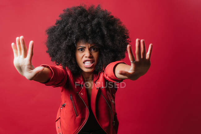 Frightened African American female with curly hair showing stop gesture on red background in studio and looking at camera — Stock Photo