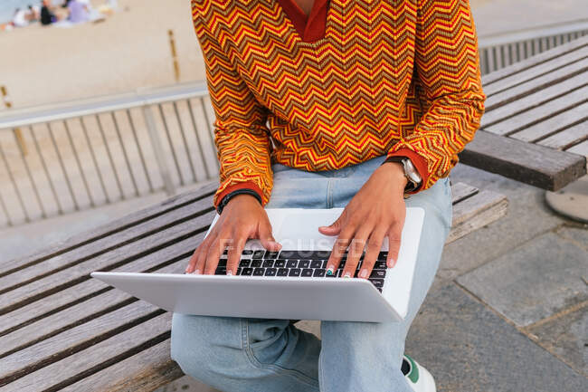 Обрізаний невпізнаваний позитивний молодий етнічний студент у стильному вбранні на клавіатурі ноутбука, сидячи на лавці біля моря на міському пляжі — стокове фото