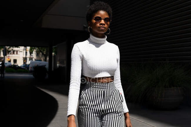 Jovem mulher negra em roupa elegante e óculos de sol enquanto caminha e olha para a câmera no dia ensolarado na rua da cidade — Fotografia de Stock