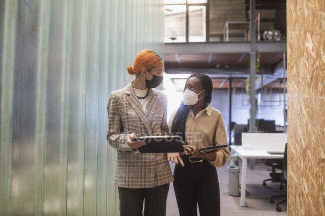 Colegas multiétnicas com tablets e máscaras protetoras em pé no espaço de co-trabalho e discutindo projeto — Fotografia de Stock