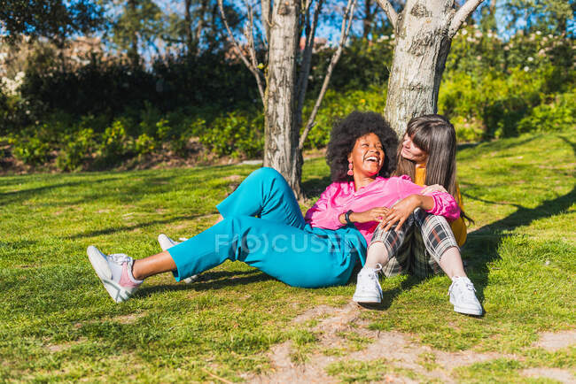Многорасовая пара гомосексуальных женщин, отдыхающих на лугу в парке, обнимаясь и веселясь в солнечный день на летних выходных — стоковое фото