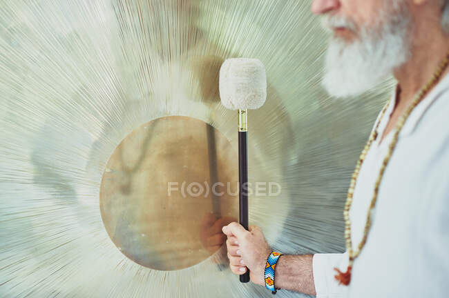 Vista lateral de la cosecha macho adulto en ropa blanca con mazo jugando gong suspendido durante la práctica espiritual - foto de stock