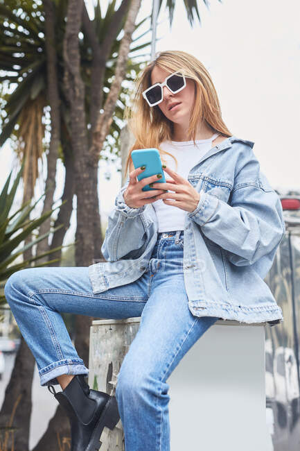 Модная женщина в джинсовой одежде сидит на улице и просматривает социальные сети на мобильном телефоне — стоковое фото