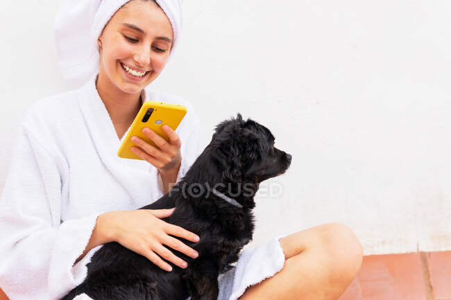 Mulher feliz em roupão de banho e toalha de estimação cão preto e celular de navegação enquanto sentado de pernas cruzadas no chão durante a rotina de cuidados com a pele — Fotografia de Stock