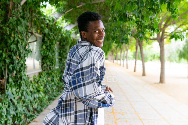 Back view of glad African American male in plaid shirt standing in city street and looking at camera over shoulder — Stock Photo