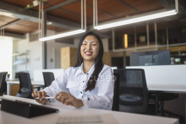 Positiva asiática empleada sentada en la mesa en el espacio de trabajo y mirando a la cámara - foto de stock
