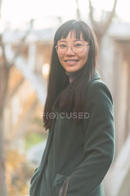 Contenu charmant asiatique femelle dans lunettes et manteau debout dans parc en automne et en regardant caméra — Photo de stock