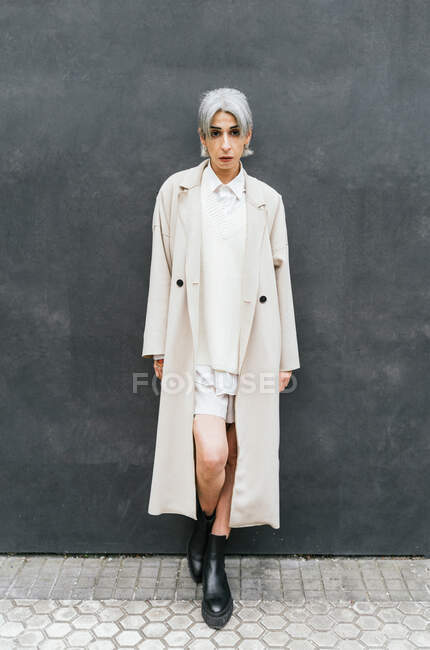 Mujer transexual seria en abrigo de moda apoyada en la pared gris mientras está de pie en la calle mirando a la cámara - foto de stock
