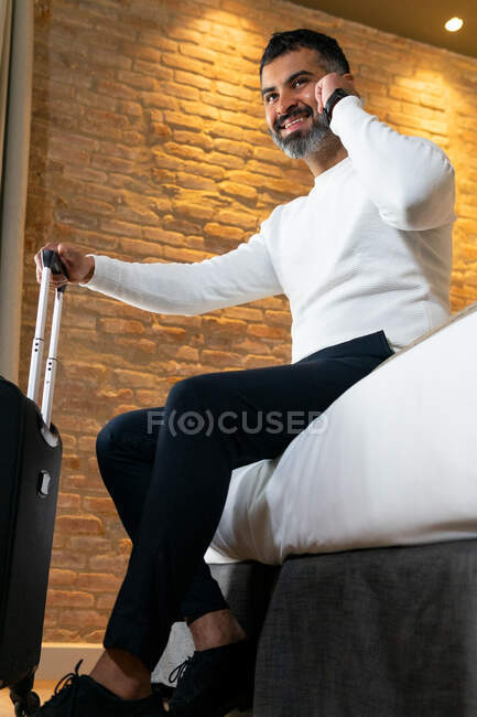Низкий угол положительного урожая этнических мужчин путешественник сидит на кровати рядом с чемоданом и говорить мобильный телефон в номере отеля — стоковое фото