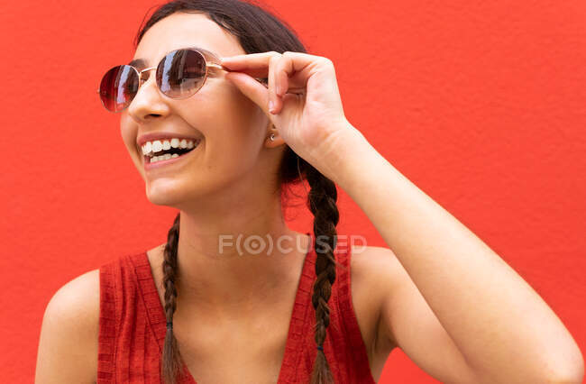 Delighted young female with pigtails putting on trendy sunglasses against red background in city street — Stock Photo
