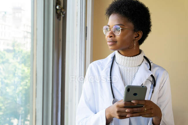 Young African American female doctor in white coat with stethoscope checking information on mobile phone while standing near window in clinic — Stock Photo