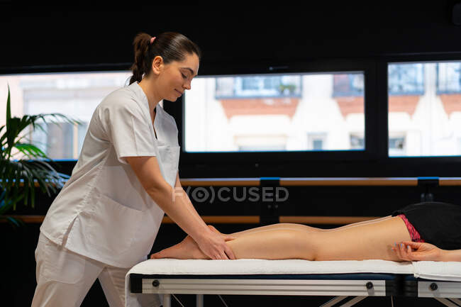 Vista laterale della massaggiatrice felice in accappatoio bianco massaggiare polpaccio di coltura paziente femminile durante la sessione di fisioterapia in clinica — Foto stock
