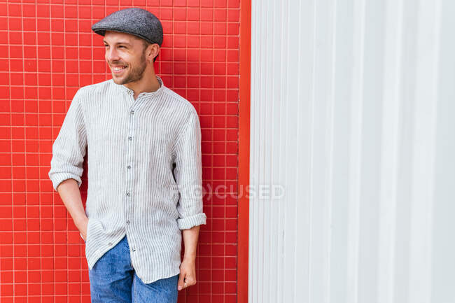 Guapo joven barbudo en camisa casual de moda y jeans y gorra de pie cerca de la pared roja y blanca y mirando hacia otro lado con sonrisa - foto de stock