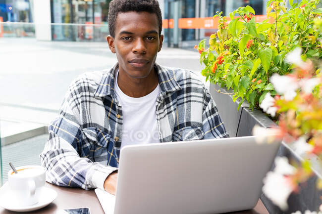 Feliz afroamericano freelancer masculino navegando y trabajando remotamente en el ordenador portátil en la cafetería al aire libre mientras está sentado mirando a la cámara en la mesa con una taza de café - foto de stock