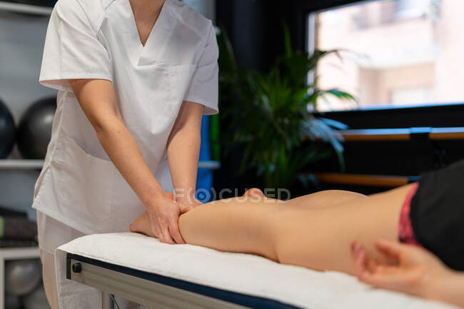 Masseuse récoltée méconnaissable en robe blanche massant le mollet d'une patiente de culture lors d'une séance de physiothérapie en clinique — Photo de stock