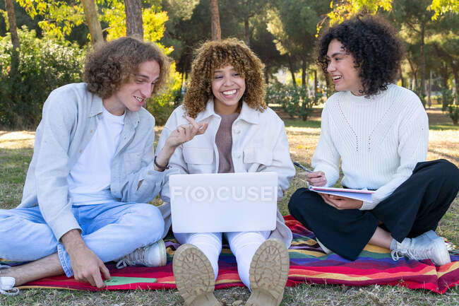 Багатоетнічний чоловік і жінки з кучерявим волоссям, сидячи на газоні в парку, використовуючи ноутбук і загальний блокнот — стокове фото