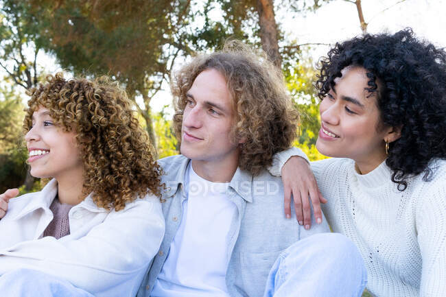 Baixo ângulo do homem abraçando diversas amigas do sexo feminino, todas com cabelo encaracolado enquanto arrepiam no parque olhando para longe — Fotografia de Stock
