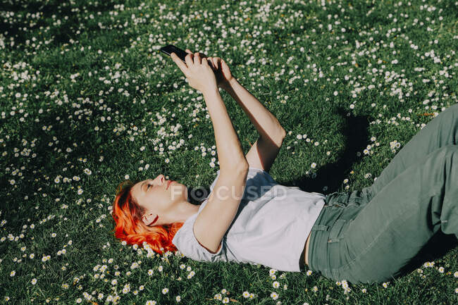 Vista lateral da fêmea ruiva encantada deitada na grama no gramado e mensagens no telefone móvel no verão — Fotografia de Stock
