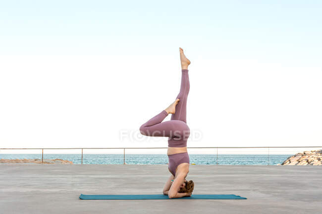Vista laterale di fit femminile fare yoga a Salamba Sirsasana mentre bilanciamento sul tappeto vicino al mare — Foto stock