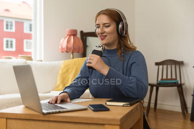 Glückliche Frau im Kopfhörer, die einen Podcast aufnimmt, während sie zu Hause mit dem Mikrofon spricht und Laptop benutzt — Stockfoto