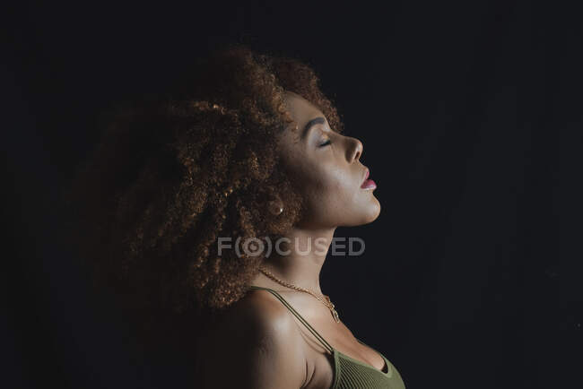 Modelo feminino afro-americano encantador com cabelo encaracolado e olhos fechados no estúdio escuro — Fotografia de Stock